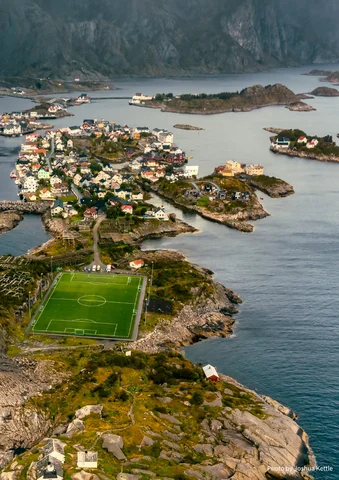 Aerial view of a coastal village with a green soccer field surrounded by water and rocky islands.