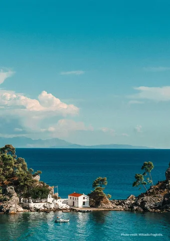 Small white house with red roof by blue sea, surrounded by rocky green cliffs under a clear sky.