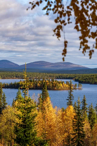 Lake surrounded by autumn trees, distant hills under a partly cloudy sky.