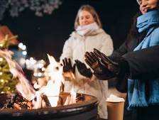 Two people warming hands over an outdoor fire pit in winter, with a Christmas tree in the background.