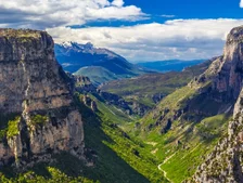 Breathtaking view of a lush valley surrounded by steep cliffs and distant snow-capped mountains under a partly cloudy sky.