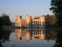 A scenic castle with turrets reflected in a calm lake, surrounded by autumnal trees under a clear blue sky.