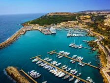 Aerial view of a vibrant marina with numerous yachts docked, surrounded by turquoise waters and lush green hills in the background.