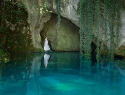 Woman in white dress stands by a cave entrance above clear blue water, surrounded by greenery.