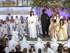 Large group of people in white robes performs a play on stage; one figure wears a bright red robe.