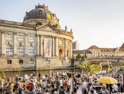 Gran multitud se reúne junto a un edificio histórico con cúpula cerca de un río en un día soleado.
