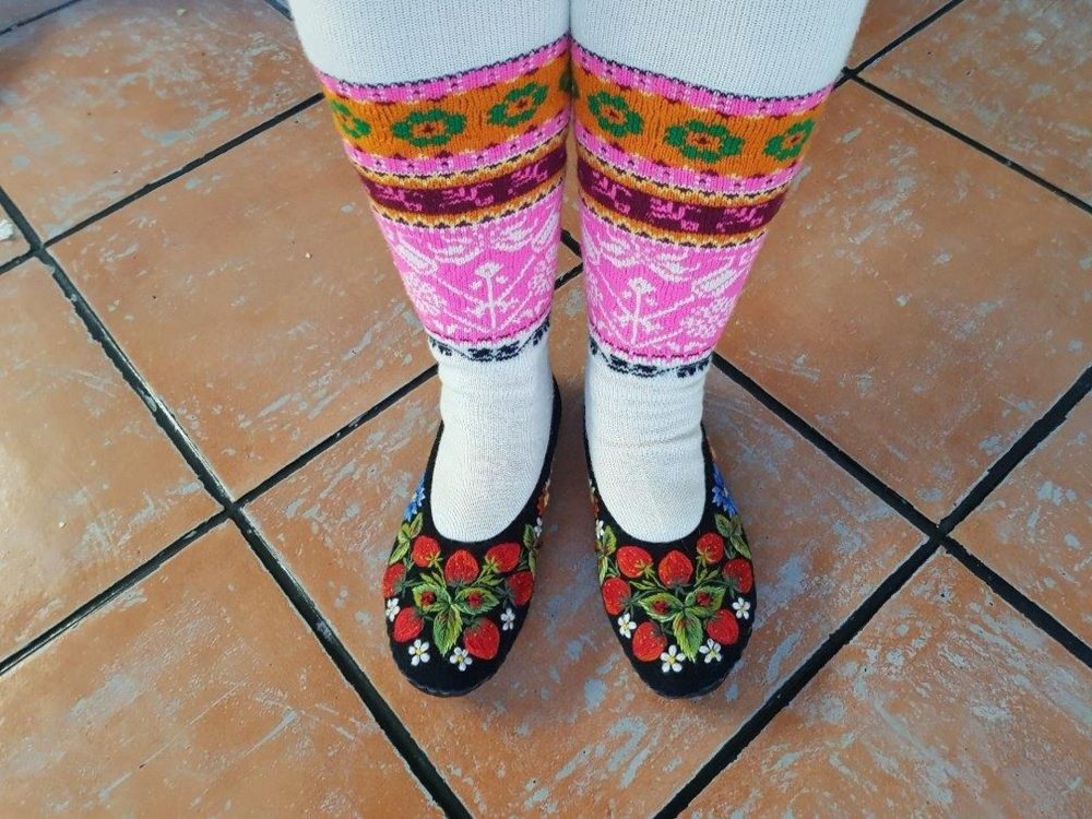 Traditional footwear featuring black embroidered flats, paired with colorful, patterned knee-high socks on tiled floor.