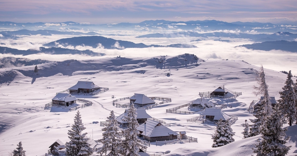 Snow-covered alpine village in Velika Planing, Slovenia, with wooden huts overlooking clouds and distant mountains.