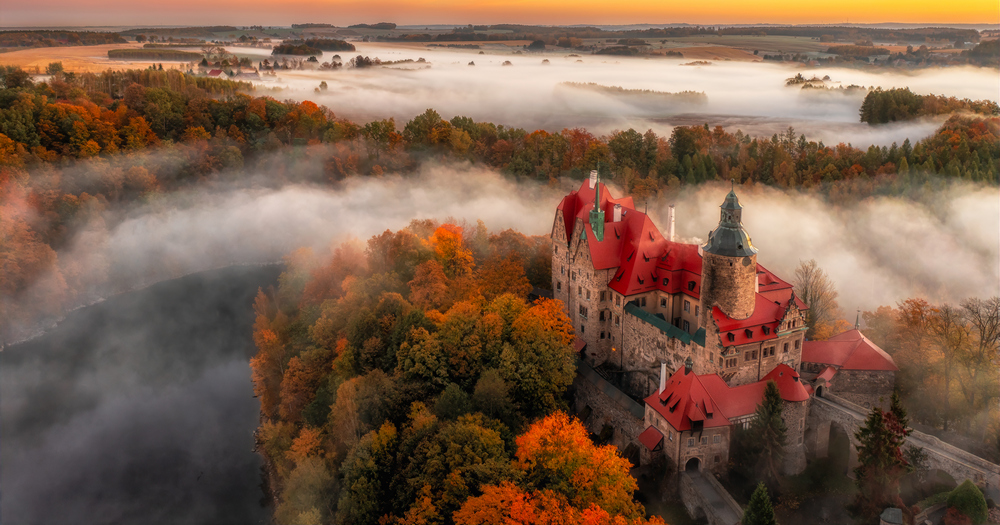 Elevated view of Czocha Castle surrounded by fall foliage in orange and green tones beneath a partly cloudy sky.