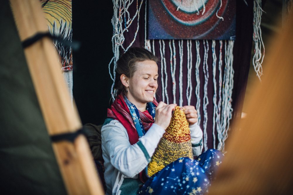 A woman smiling while knitting a colorful piece, surrounded by handmade decorations in a cozy craft setting.