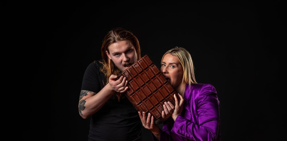 A man and woman hold a large chocolate bar, smiling and positioned against a dark background. The man has long hair and tattoos; the woman wears a purple blazer.