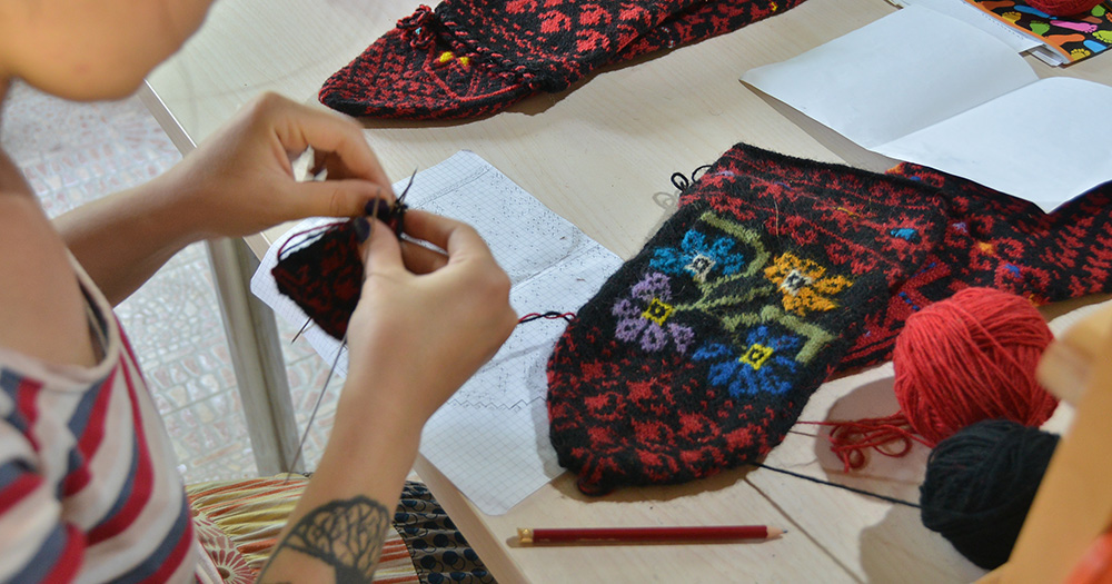 A person crafts colorful socks, surrounded by yarn and sketches, focusing on intricate needlework.