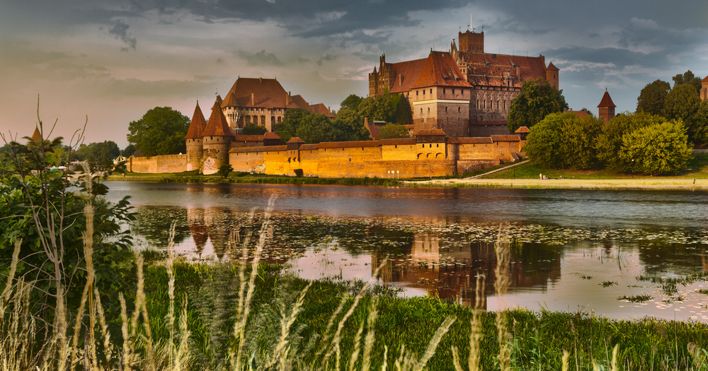 Marlborough Castle seen across a river, surrounded by lush green landscape and reflected in the water beneath clouds.