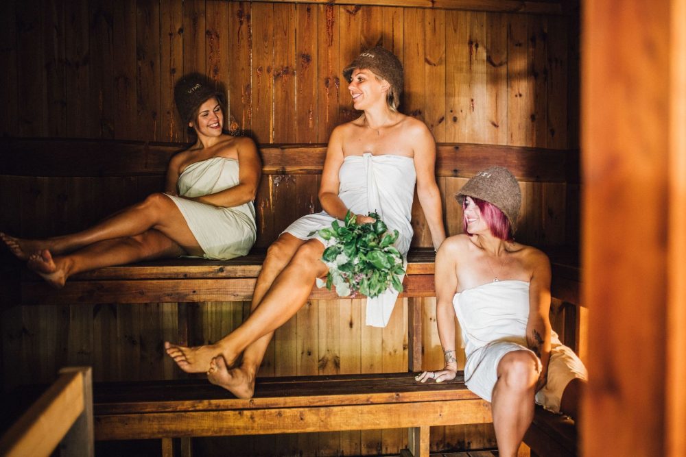 Three women in towels relax in a wooden sauna, with one holding a bundle of green leaves surrounded by a warm, rustic atmosphere.