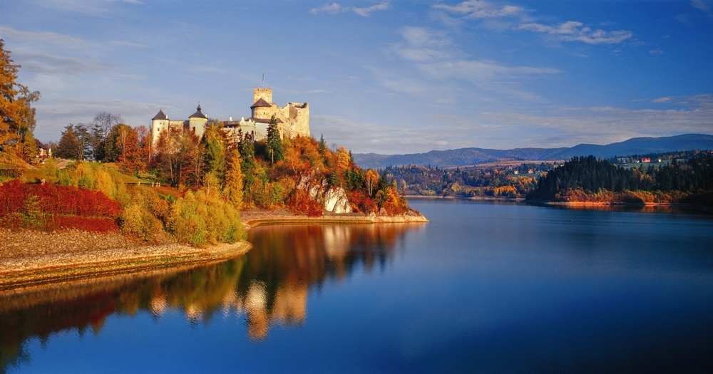 Autumn landscape at Niedzica Castle with colorful trees in green, orange, and red, mountainous backdrop, and a river winding below the hill beneath a clear sky.