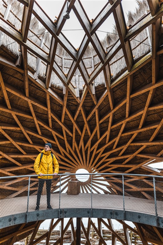 A person in a yellow jacket stands on a circular balcony beneath a striking wooden star-patterned ceiling with an opening at the center.