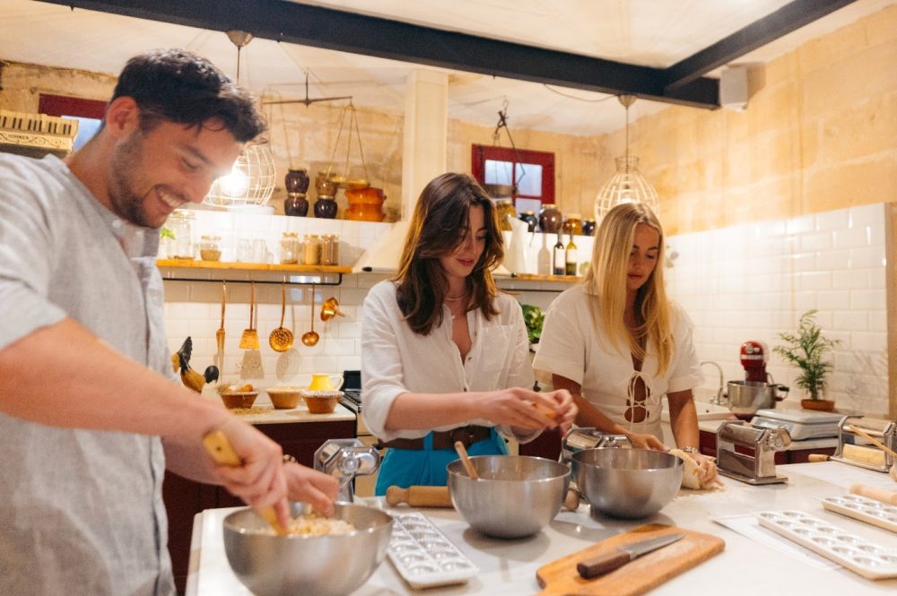 Three people are engaged in cooking Maltese bread, working together at a marble kitchen counter with bowls and baking utensils.