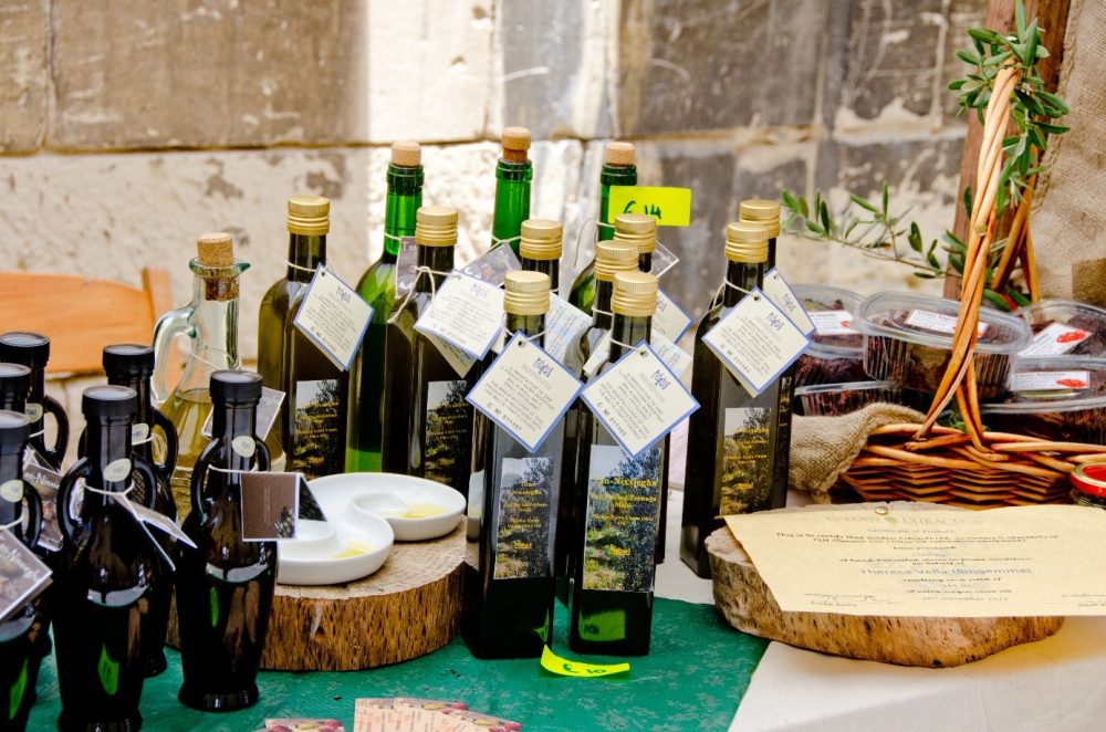 A display of various olive oil bottles with tags, alongside small dishware and a basket of preserves on a green tablecloth.