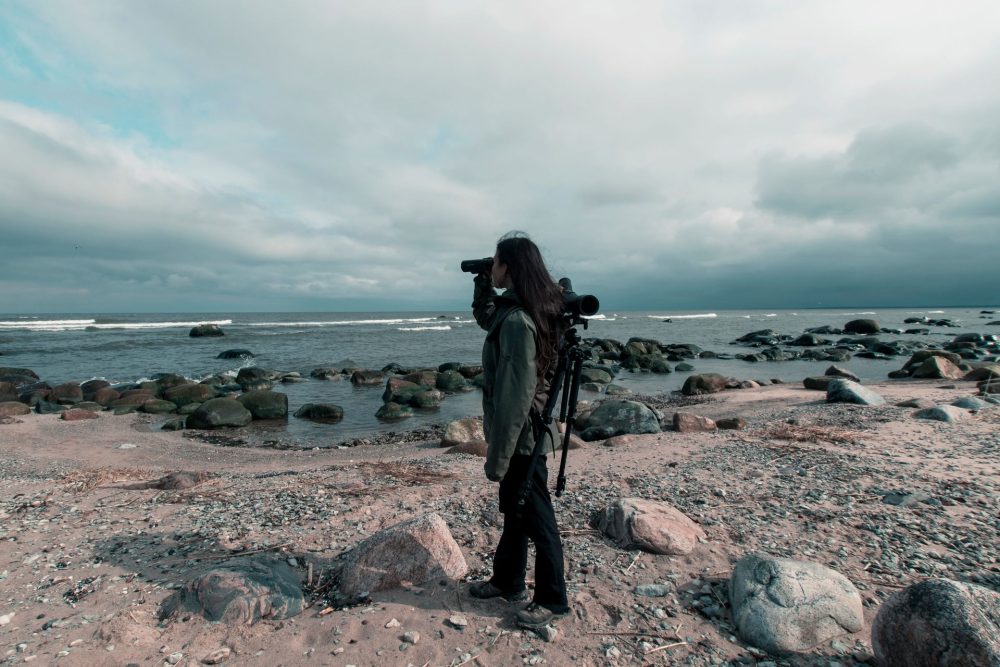 Person with binoculars beside a camera tripod on a rocky shoreline under cloudy skies.