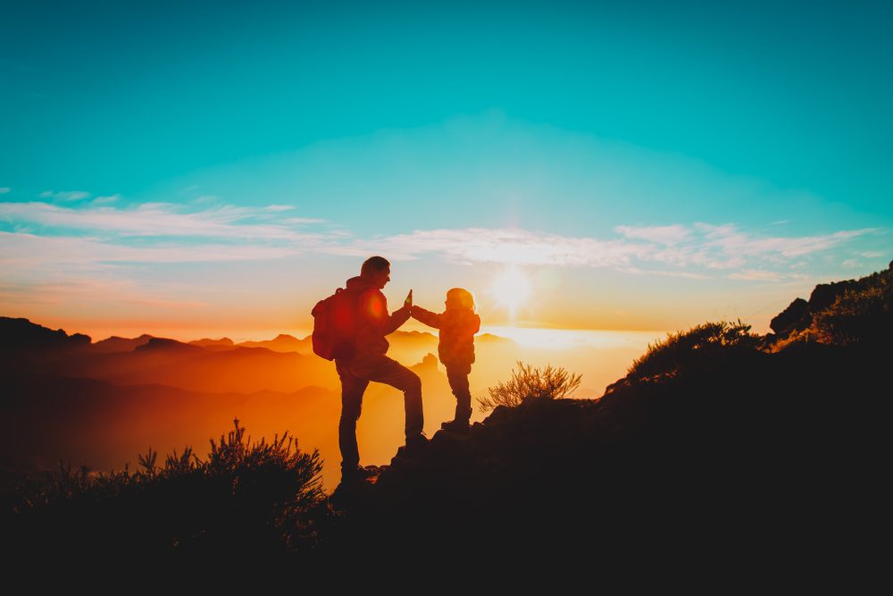 A silhouetted adult and child high-five on a mountain summit at sunset, with dramatic orange light casting over distant peaks and a vibrant blue sky above.