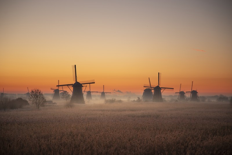 Sunset view of the windmills with clouds between them, painted in orange, yellow, and blue hues, with dry brown grass in the foreground.