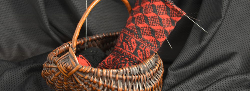 A woven basket holds a partially completed red and black patterned textile piece with sewing needles, against a dark fabric backdrop.