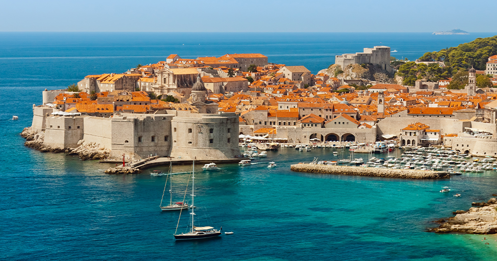 Aerial view of the medieval city walls, historic port, and terracotta roofs of Dubrovnik, Croatia, overlooking the turquoise Adriatic Sea.