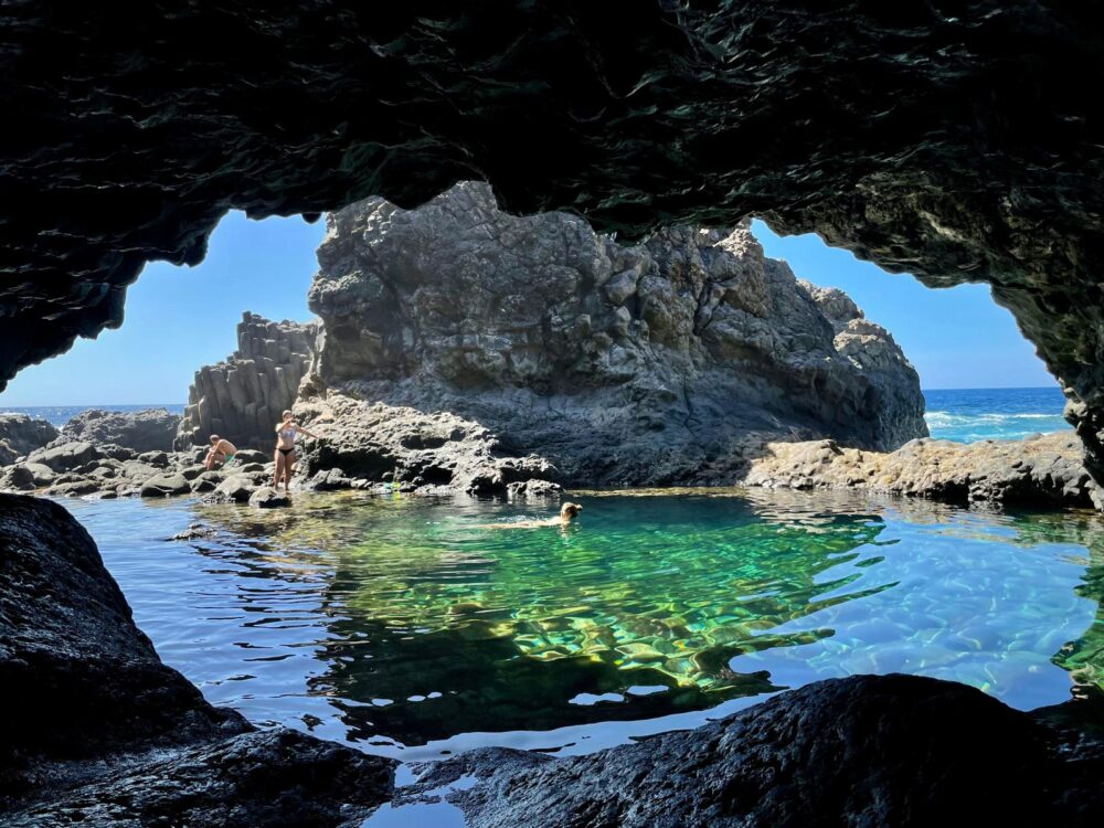 View from a cave of a clear tidal pool surrounded by rocks, with a bright blue sky above.