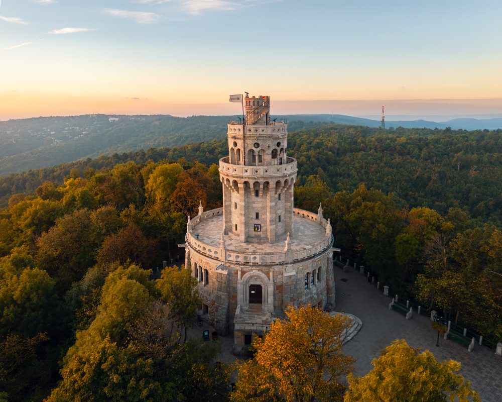 Majestic stone tower of the Erzsébet Lookout surrounded by a the Normafa park with autumn trees, located atop a hill with panoramic views of the surrounding landscape.