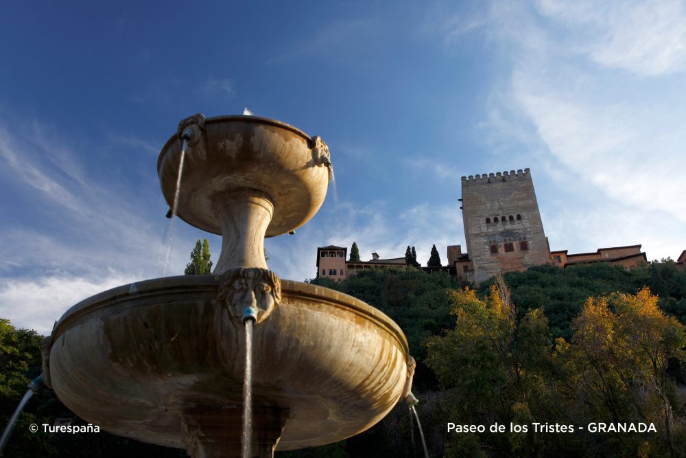 Photo taken from below featuring a fountain in the foreground with water flowing from its sculptures, the Paseo de los Tristes behind it, the castle in the background, green trees, and a blue sky above, illuminated by sunlight.