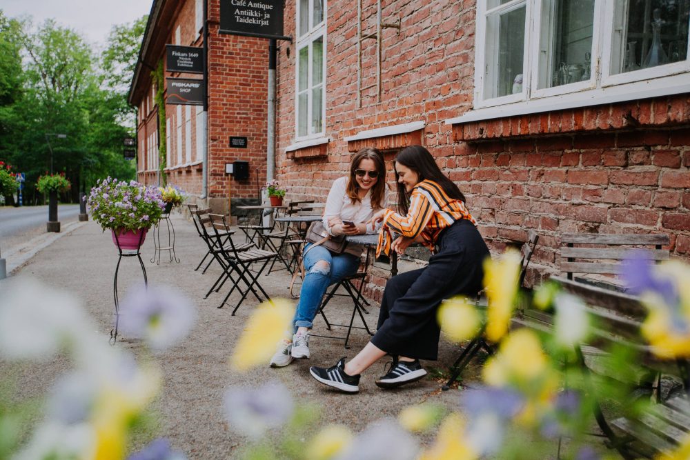 Friends sitting in a café in Southern Finland, smiling and looking at a phone, surrounded by flowers with green trees in the background.