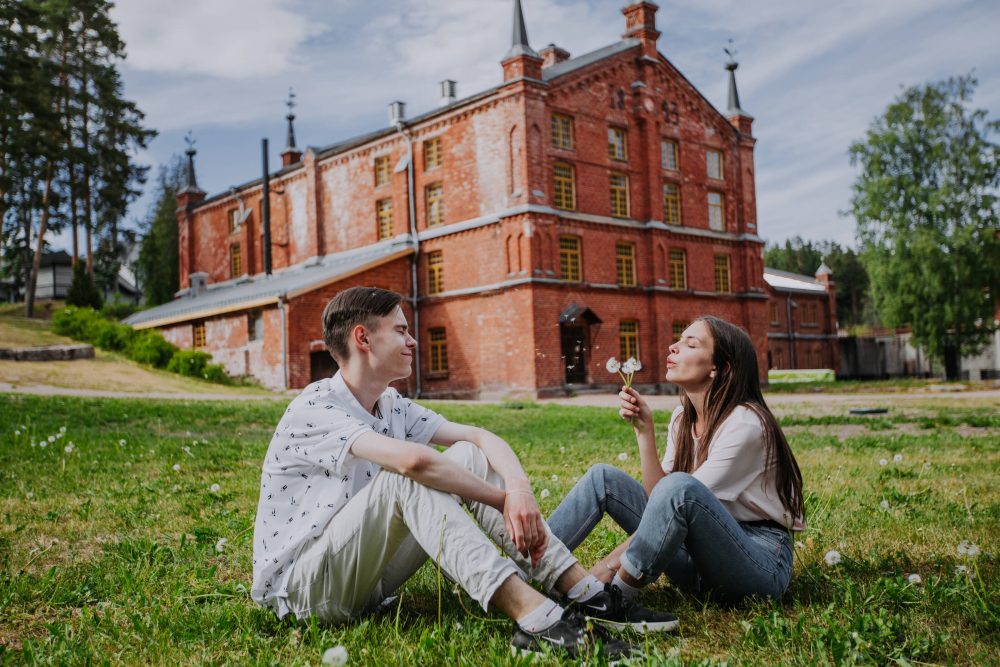 Couple sitting in front of the Kouvola board mill, a UNESCO World Heritage site, blowing a dandelion on a summer day, surrounded by green scenery.