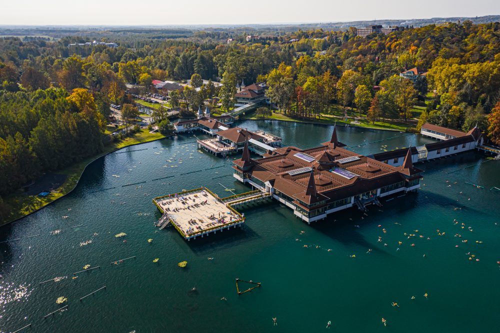 Aerial view of the Hévíz Thermal Lake and its surrounding spa buildings, set amidst lush green foliage and autumn-coloured trees.