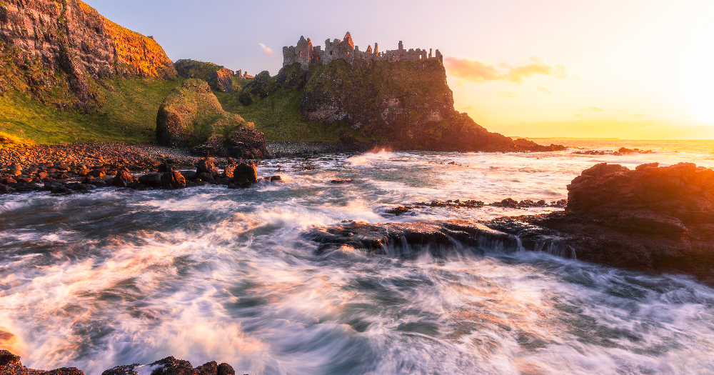 Dunluce Castle perched dramatically on a rugged cliff in County Antrim, Northern Ireland, with the Atlantic Ocean crashing below and a vibrant sunset sky above.