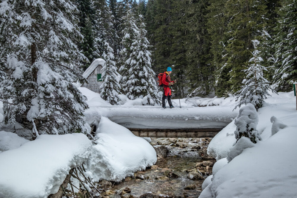 A person wearing red hiking gear and a blue hat crosses a snowy wooden bridge surrounded by a winter forest, with a small cabin visible in the background.