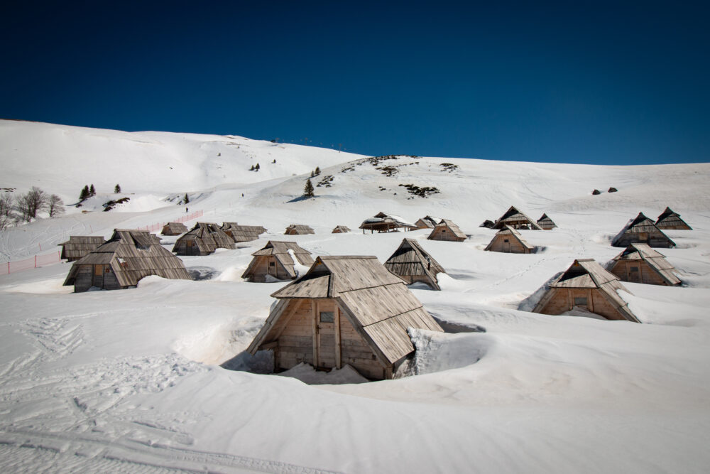 A village of traditional wooden huts partially buried in snow on a pristine white mountainside, under a clear blue sky.