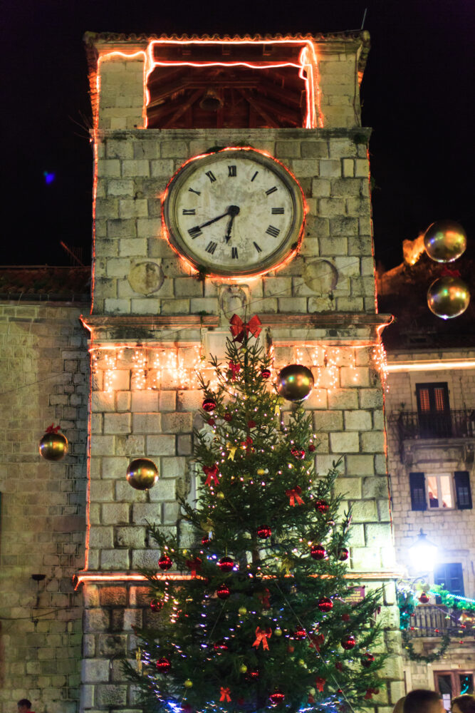 A clock tower decorated with lights. In front of it, a Christmas tree.