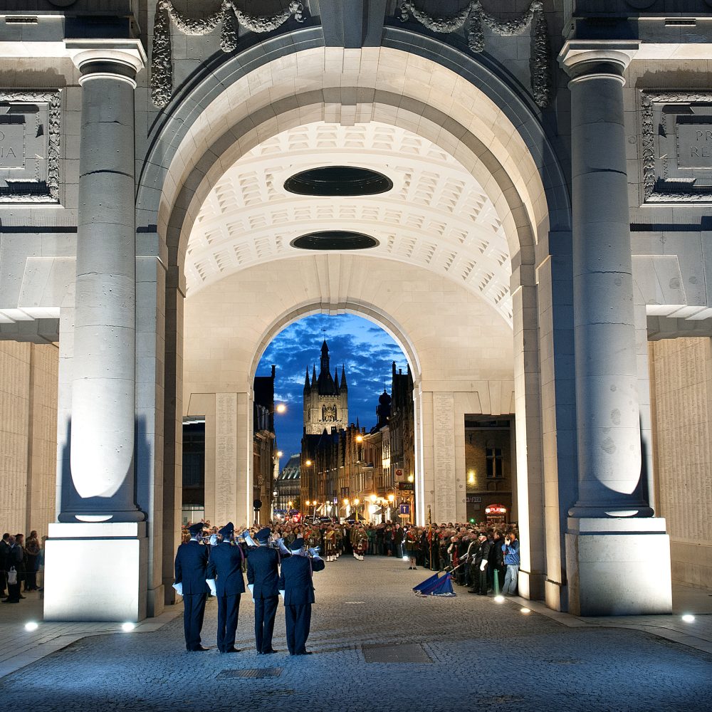 Night scene of soldiers playing trumpets during The Last Post ceremony, illuminated by lights, with a cathedral in the background and people watching.