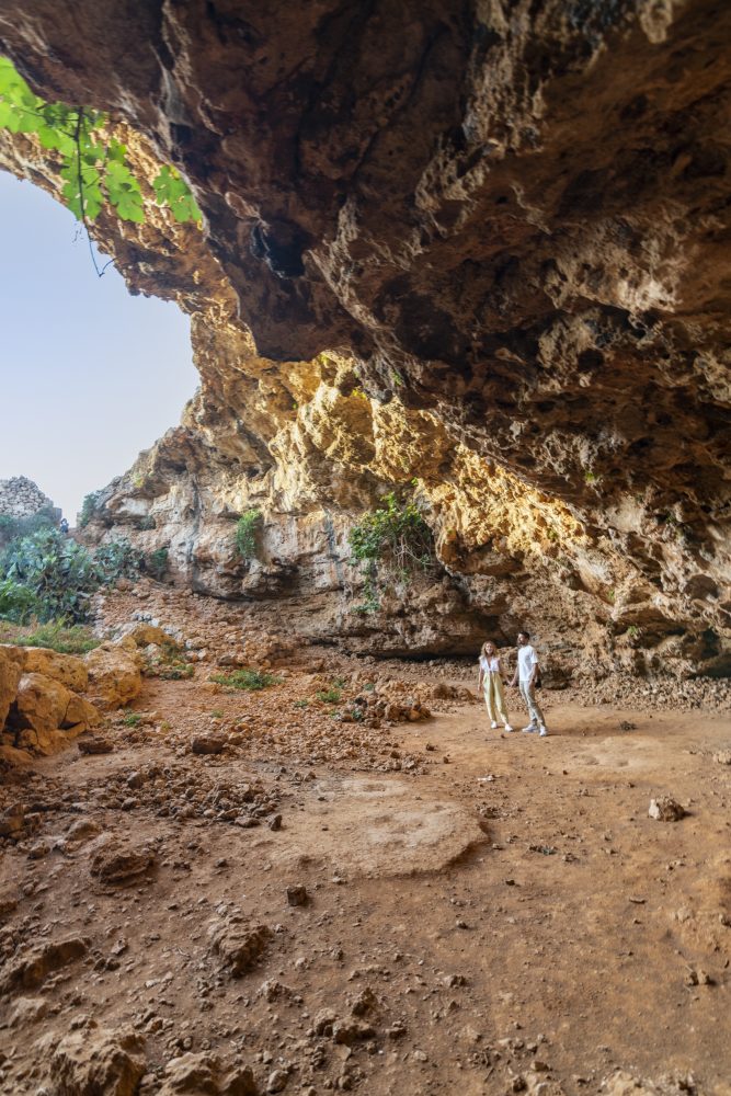 A couple explores a rocky cave, surrounded by textured stone walls and sparse greenery, under a bright sky.