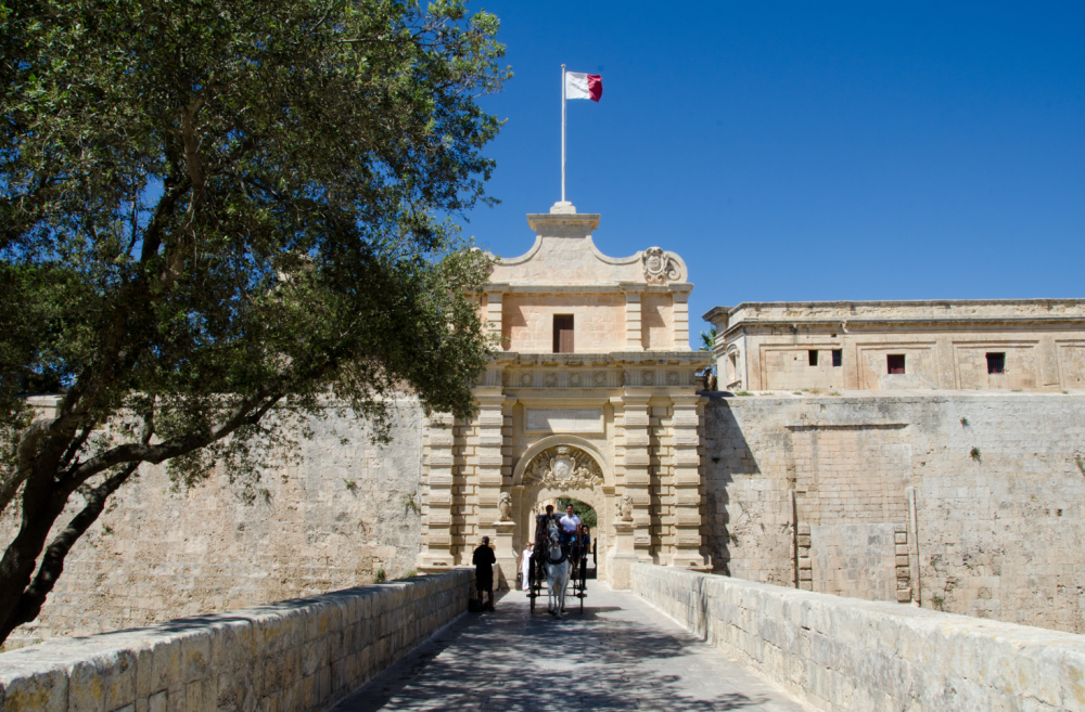 Historic gate of Mdina with a flag overhead, visitors passing through, and a tree casting shade over the bridge.