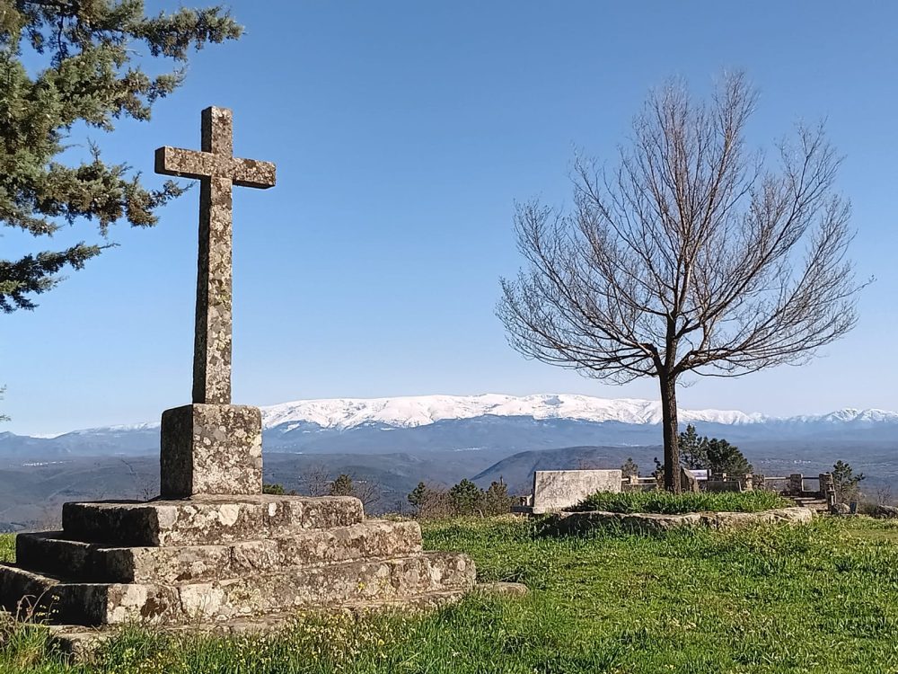 Stone cross and bare tree overlooking snowy mountains under a clear blue sky.