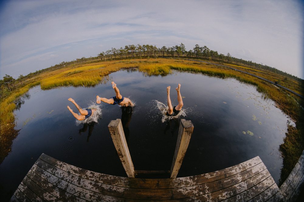 Three people diving into a calm, reflective pond surrounded by lush greenery and a clear blue sky, splashing water dramatically.