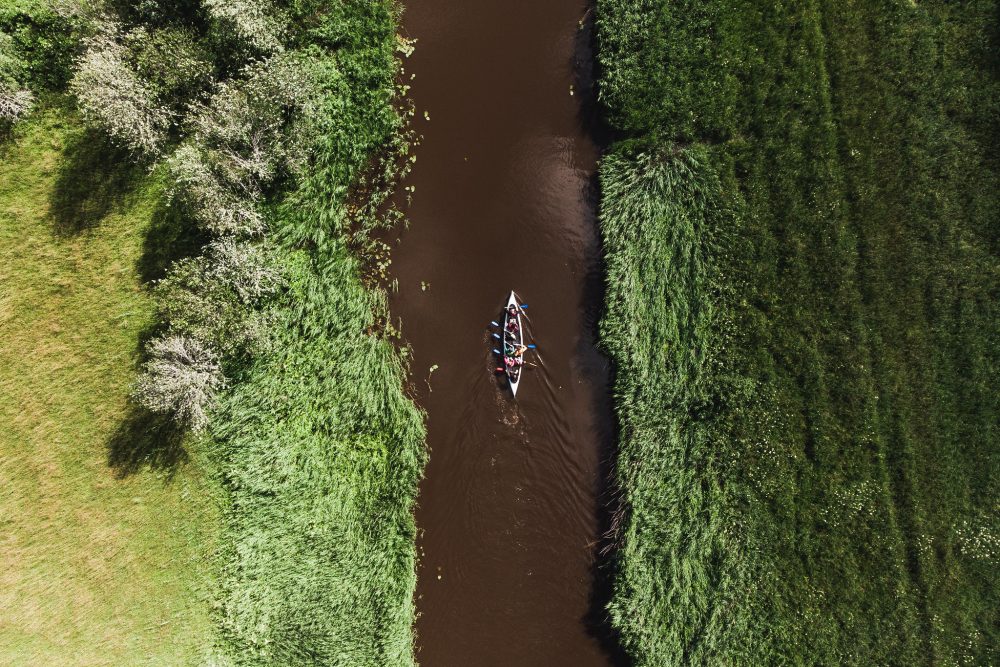 Aerial view of a canoe with several paddlers navigating a narrow, winding river bordered by lush greenery and wildflowers.