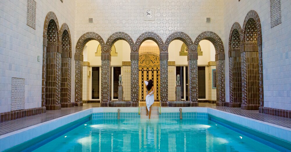 A woman by an indoor swimming pool in the Kaiser-Friedrich-Therme in Germany, a spa with ornate arches and mosaic tiles.