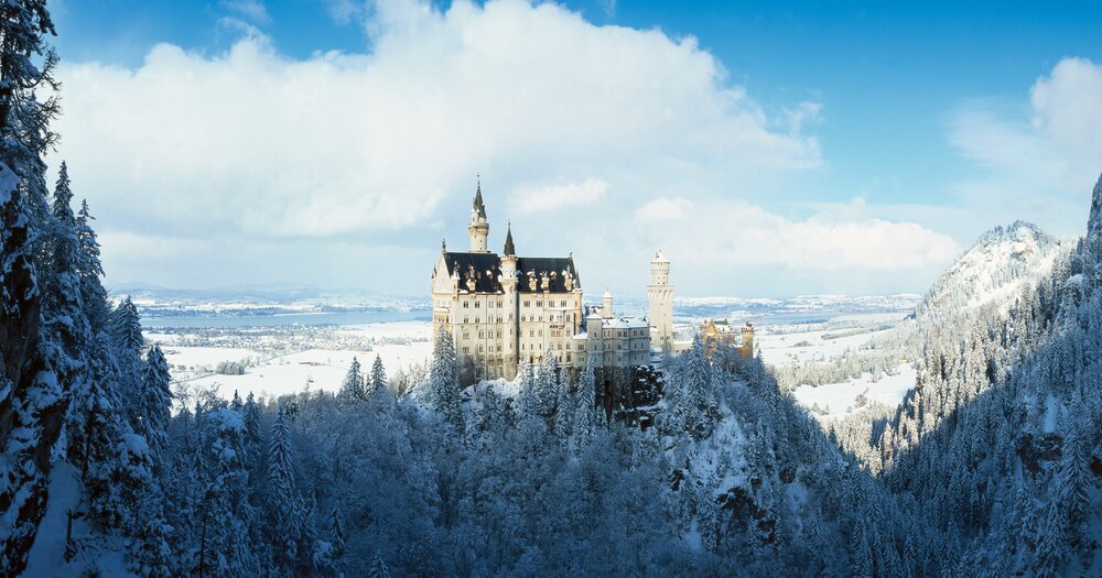 A panoramic view of the snow-covered castle Neuschwanstein Castle in Schwangau in Germany, amidst pine forests with a blue sky overhead.