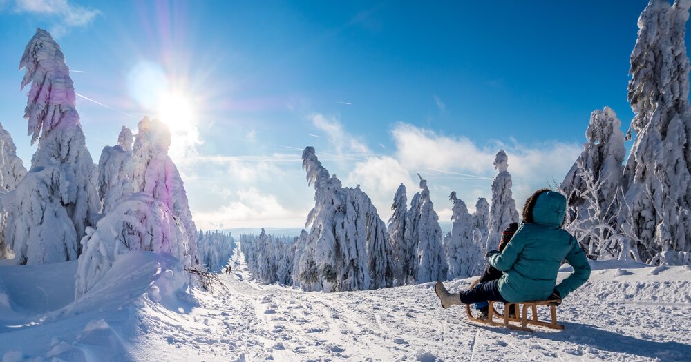 A mother and a child on a sled looking at snowy trees and mountains under a bright sun in Erzgebirge, Germany.