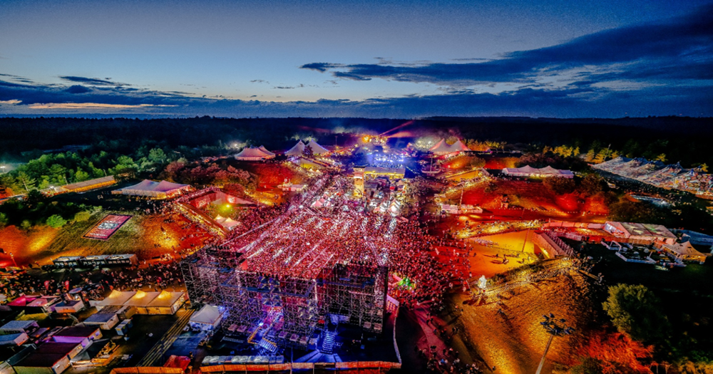 从空中俯瞰，一个规模庞大、人潮涌动的户外音乐节，舞台灯光璀璨灯光，多个大型升降台结构林立。