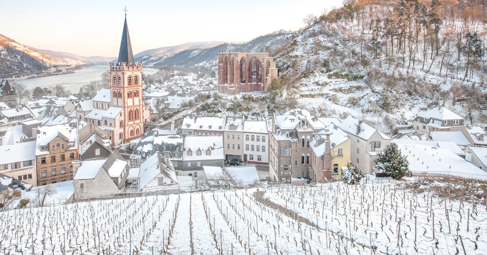 A snowy view of Bacharach im Mittelrheinta with a vineyard, St. Peter`s Church, Stahleck Castle Sankt-Johanner-Markt and buildings by a river.