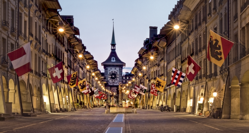 Historic street in Bern, Switzerland, decorated with Swiss and cantonal flags, leading to the iconic Zytglogge clock tower at dusk.