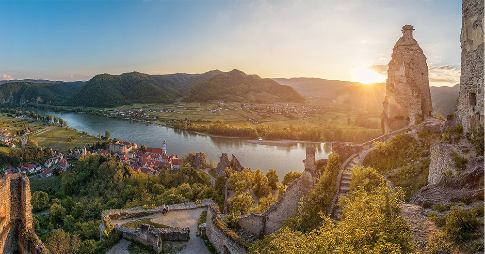 A high-angle panoramic sunset view of the Danube River valley, showing the red-roofed village of Dürnstein nestled by the water, framed by the sun-drenched stone ruins of Dürnstein Castle in the foreground.